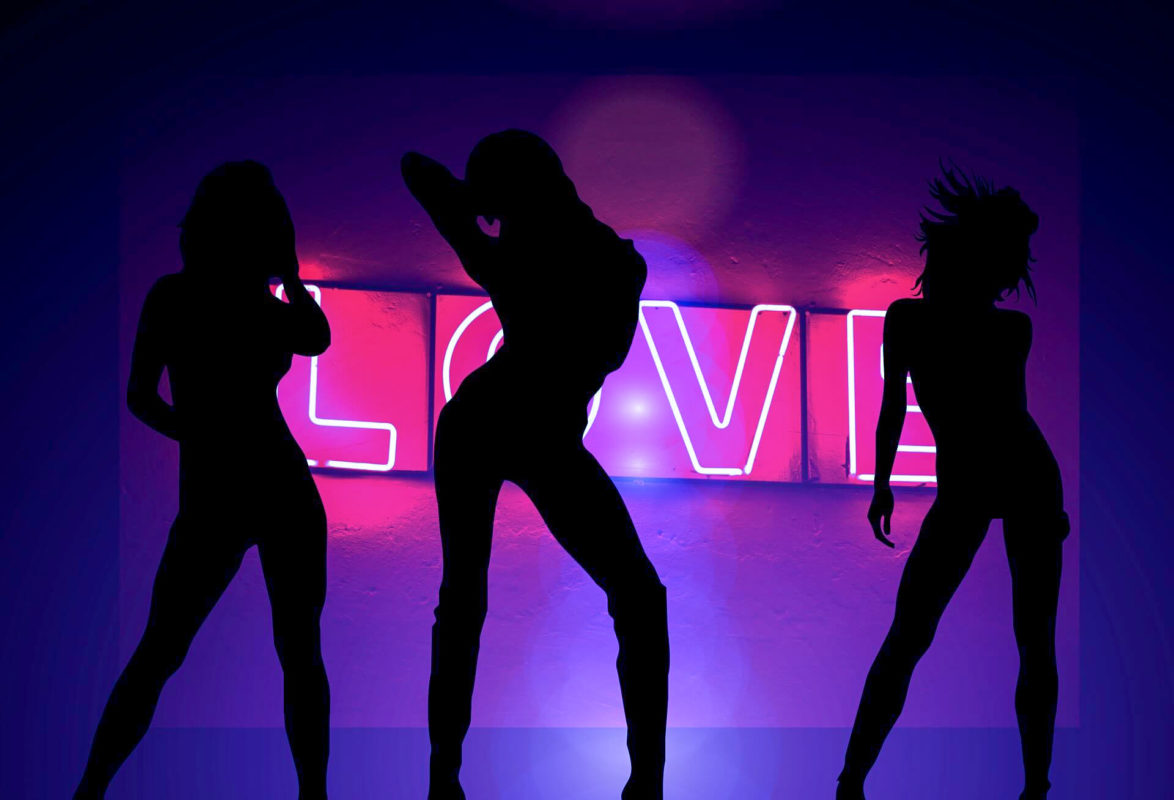 Drei Frauen-Silhouetten tanzen vor einem pinken Love-Neon-Schriftzug. 